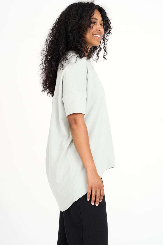 Side view of woman wearing light sage green Tencel short sleeve slouchy top with back pleat detail, made by Canadian loungewear label Advika. 